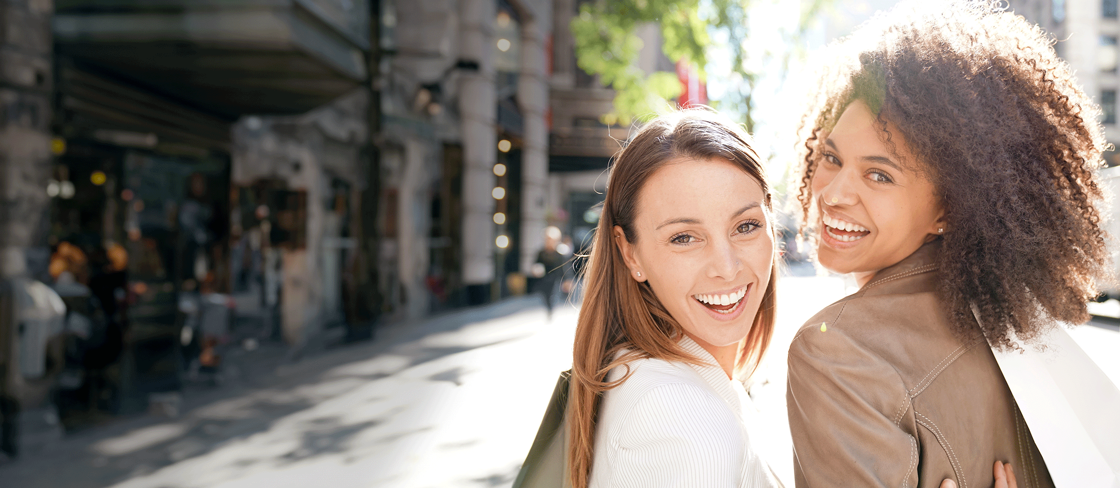 Twee lachende vrouwen in een zonnige stadsomgeving, genietend van winkelen en stedelijke lifestyle