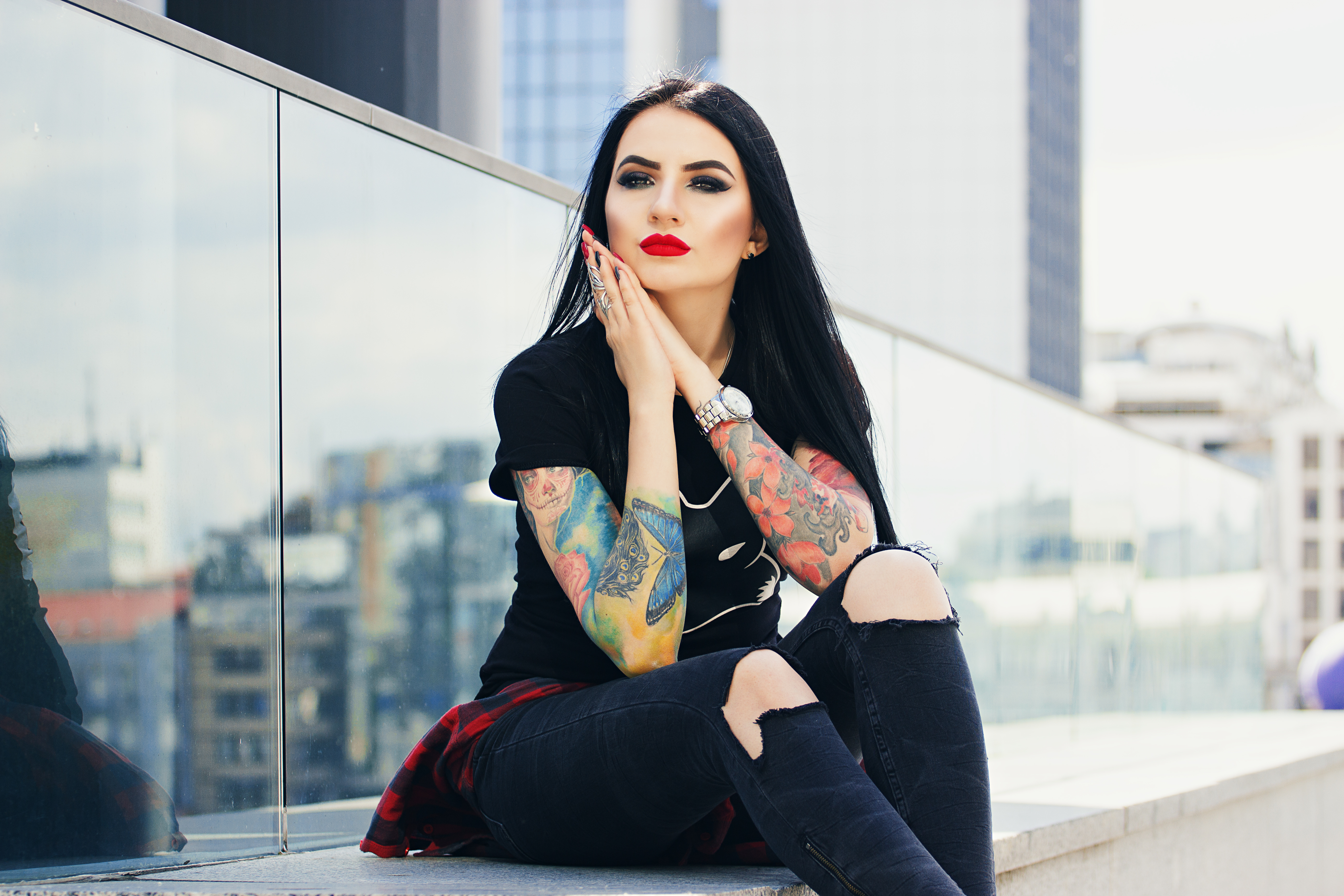 Vrouw met lange zwarte haren en kleurrijke tatoeages, zittend buiten tegen een glazen achtergrond.